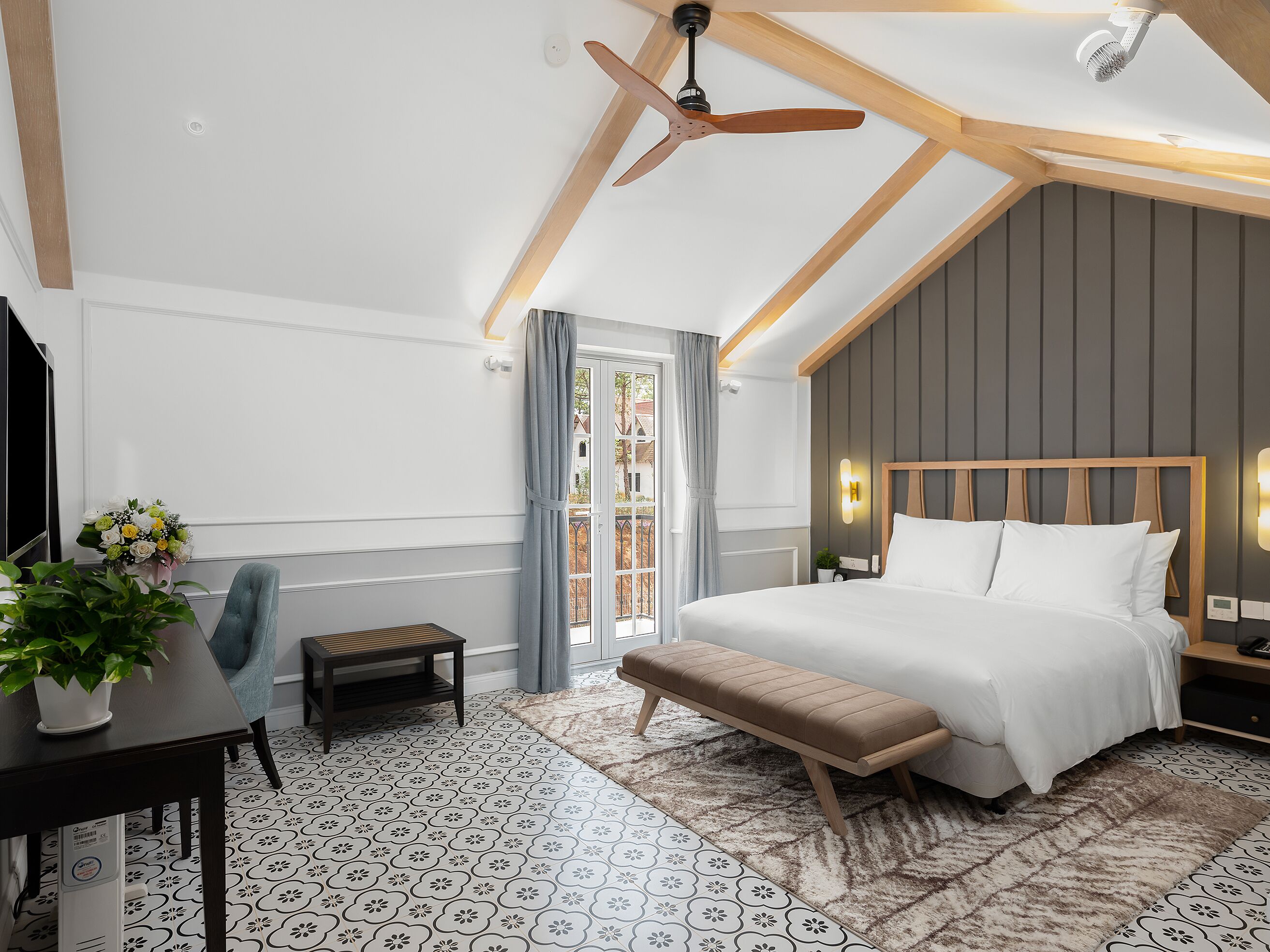 A bedroom with a bed, a bench, a desk with a plant, a TV, and a ceiling fan.