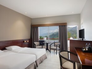 A hotel room with two beds, a desk with a TV, a dining area with chairs, and a large window with a mountain view. - Niagara Hotel Lake Toba Resorts (Parapat)