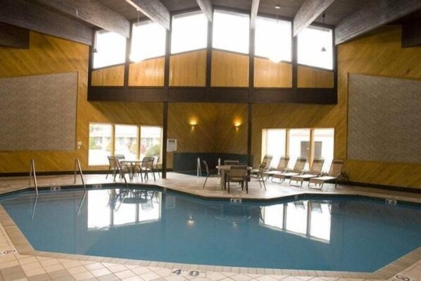 An indoor swimming pool with a tiled floor, lounge chairs, and a high ceiling with wooden beams.