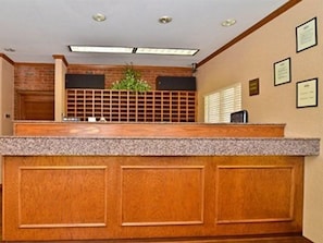 A reception desk with a wooden finish, a plant, and framed certificates on the wall. - Suburban Studios Stuart Near Federal Hwy 1 (Stuart)