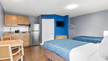 A compact room with a kitchenette, a bed with patterned bedding, a TV mounted on the wall, and a wooden chair.