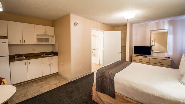 A compact hotel room with a kitchenette, a bed, a bedside table, a mirror, and a door leading to a bathroom. - Suburban Studios Stuart Near Federal Hwy 1 (Stuart)