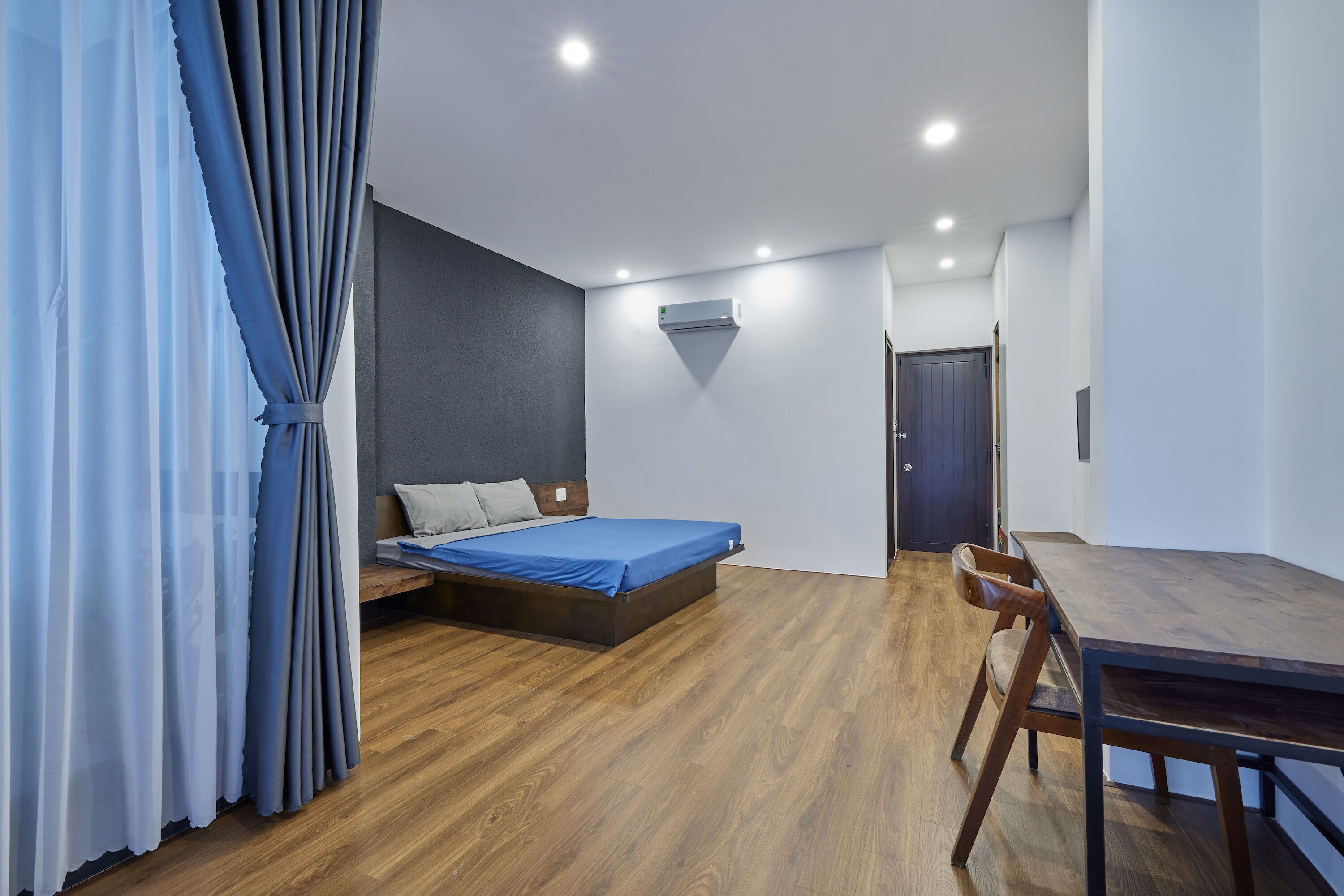 A modern bedroom with a wooden floor, a bed with blue bedding, a desk, and a chair.