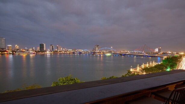 A waterfront view with a modern bridge, city skyline, and a bar area with seating. - The Garden Capsule Hotel (Da Nang)