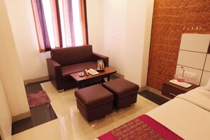 A hotel room with a sofa, ottomans, a small table with a kettle, a tray with a bottle and glasses, and a bed with a patterned bedspread. - Hotel Sky View (New Delhi)
