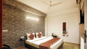 Une chambre d’hôtel avec un mur d’accent en briques, un lit avec du linge de lit blanc, une table de chevet, un bureau et un ventilateur de plafond.