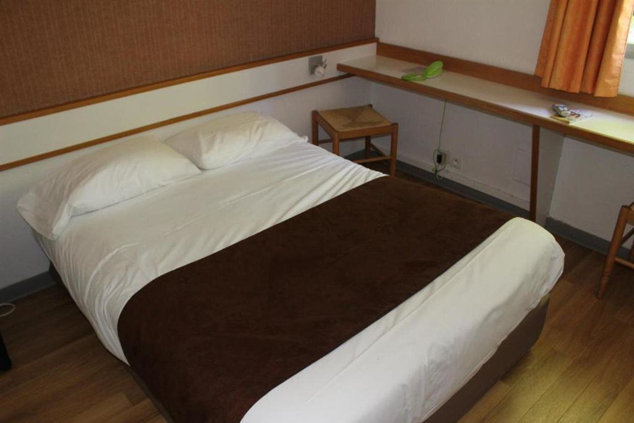 A single bed with a brown blanket, a wooden nightstand, a small table, and a window with curtains.