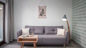 A grey sofa with two pillows, a wooden side table with books, a hanging botanical print, and a floor lamp. - Hotel Sonnenhof (Neuweiler)