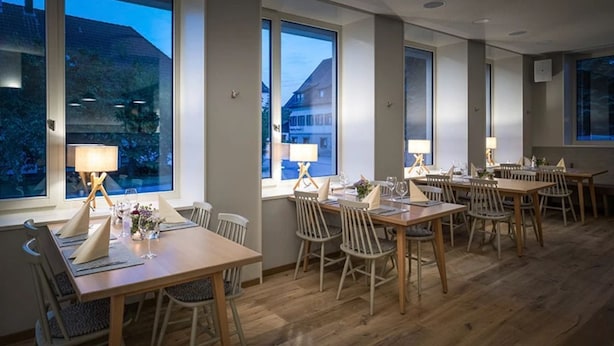 A dining area with multiple tables and chairs, large windows, and lamps on stands. - Hotel Sonnenhof (Neuweiler)