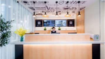 A reception desk with a light wood finish, a large window with blinds, and a wall-mounted light fixture above.