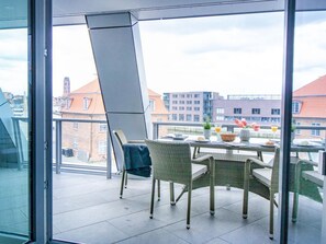 A balcony with wicker chairs and a table set for breakfast, overlooking a cityscape with buildings and a clear sky.