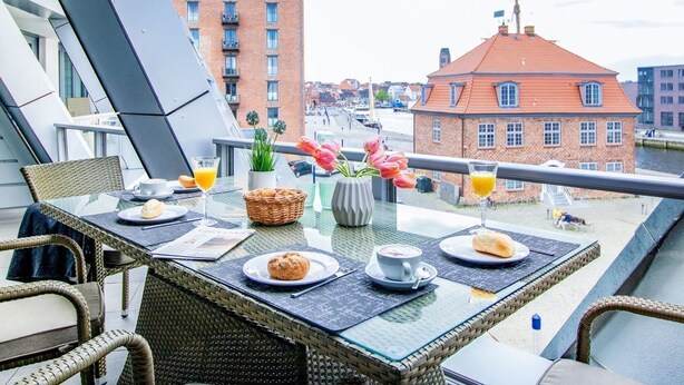 Un balcón con mobiliario de mimbre, una mesa dispuesta con alimentos para el desayuno y vistas de la ciudad.