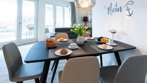 Comedor con una mesa dispuesta para el desayuno, que incluye pastelería y café, en una habitación moderna con ventanas grandes y una araña.