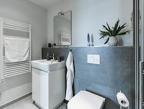 Une salle de bain moderne avec un lavabo blanc, un miroir et des toilettes. On y trouve un porte-serviettes avec des serviettes, une plante en pot et un espace douche doté d’une porte en verre.