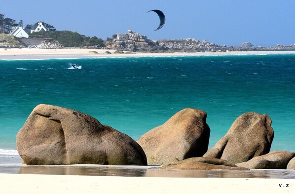 A beach with large rocks in the foreground, a clear blue sea, and a distant view of a coastal building.
