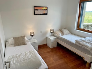 A bedroom with two single beds, a nightstand with a lamp, a small table, and a window with a view of the outdoors.