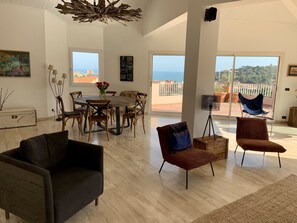 Un vaste espace de vie comprenant une table à manger en bois, des sièges confortables et de grandes fenêtres offrant une vue sur la mer.