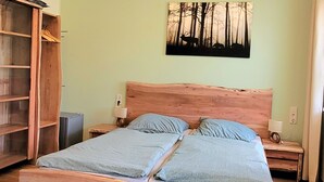 Een kamer met een houten bed, twee kussens, een nachtkastje met een lamp, een kledingkast en een schilderij van bomen aan de muur.