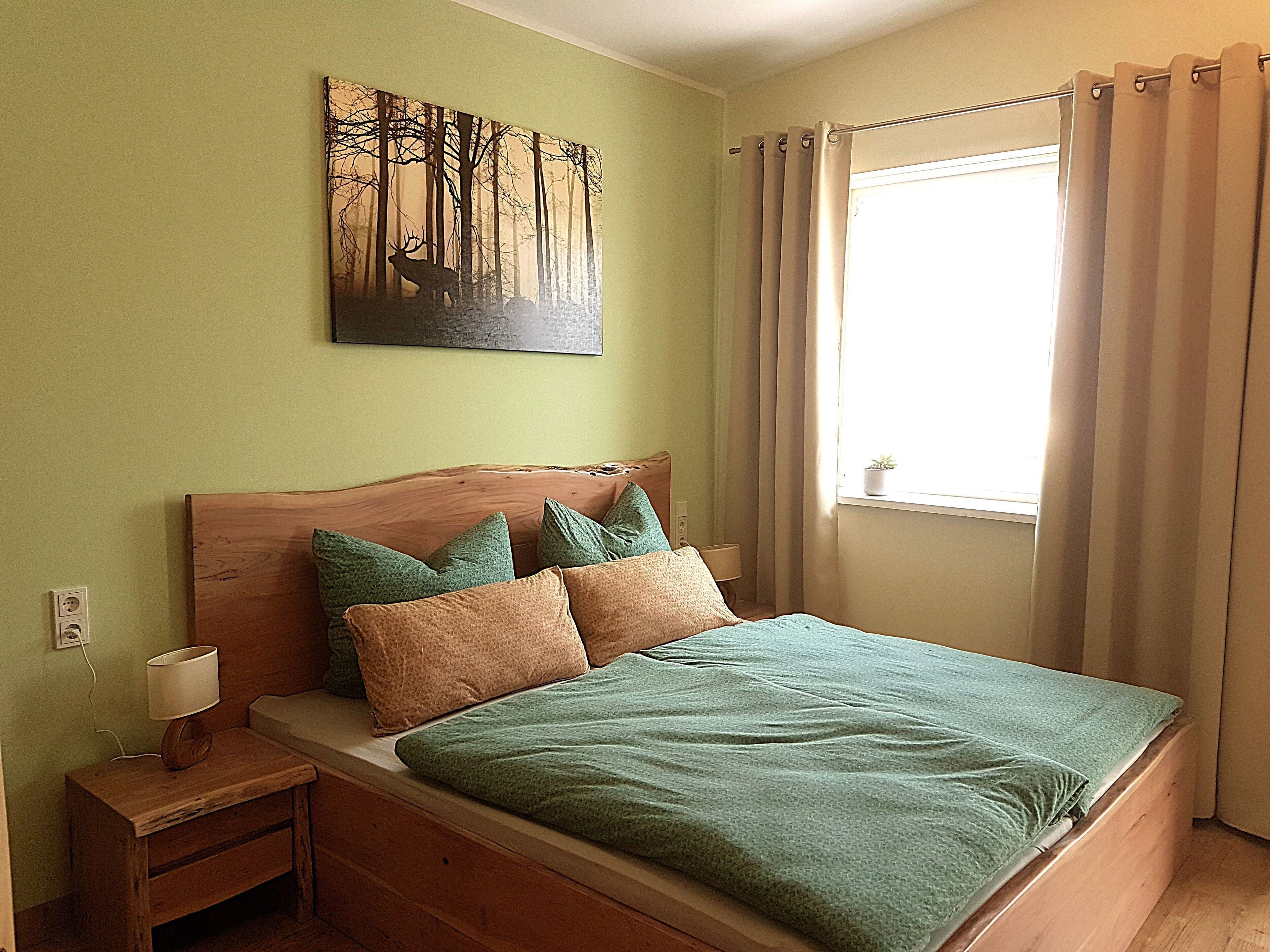 Una cama con cabecera de madera, almohadas verdes y beige, una colcha verde, una mesita de noche con una lámpara y un cuadro de un bosque en la pared.