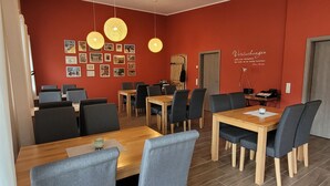 Uma área de refeições com mesas e cadeiras de madeira, uma parede vermelha com quadros e luminárias pendentes.