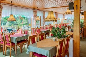 A restaurant with multiple tables and chairs, wooden ceiling, and large windows with curtains.