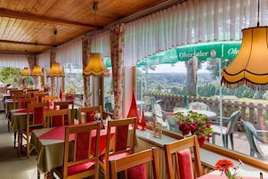A restaurant with wooden tables and chairs, large windows with curtains, and outdoor seating visible through the windows.