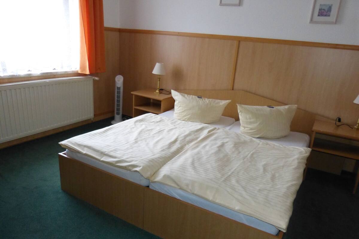 A hotel room with a bed, two bedside tables, a lamp, a radiator, and a window with curtains.