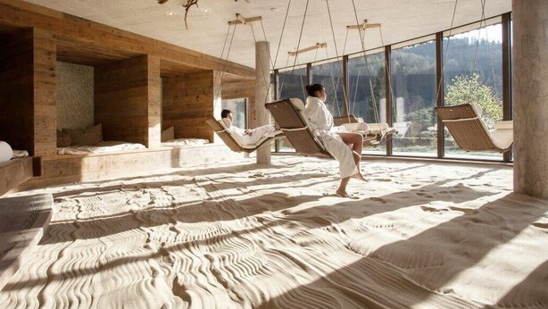 A room with wooden walls and large windows, featuring a bed, a hanging chair, and a massage table. - Feuriger Tatzlwurm Berghotel & Spa BW Signature (Oberaudorf)