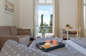 Ein Hotelzimmer mit einer Couch, einem Bett mit einem Frühstücksbrettchen, einem Tisch mit einem Schachbrett und einem Balkon mit Blick auf Bäume.
