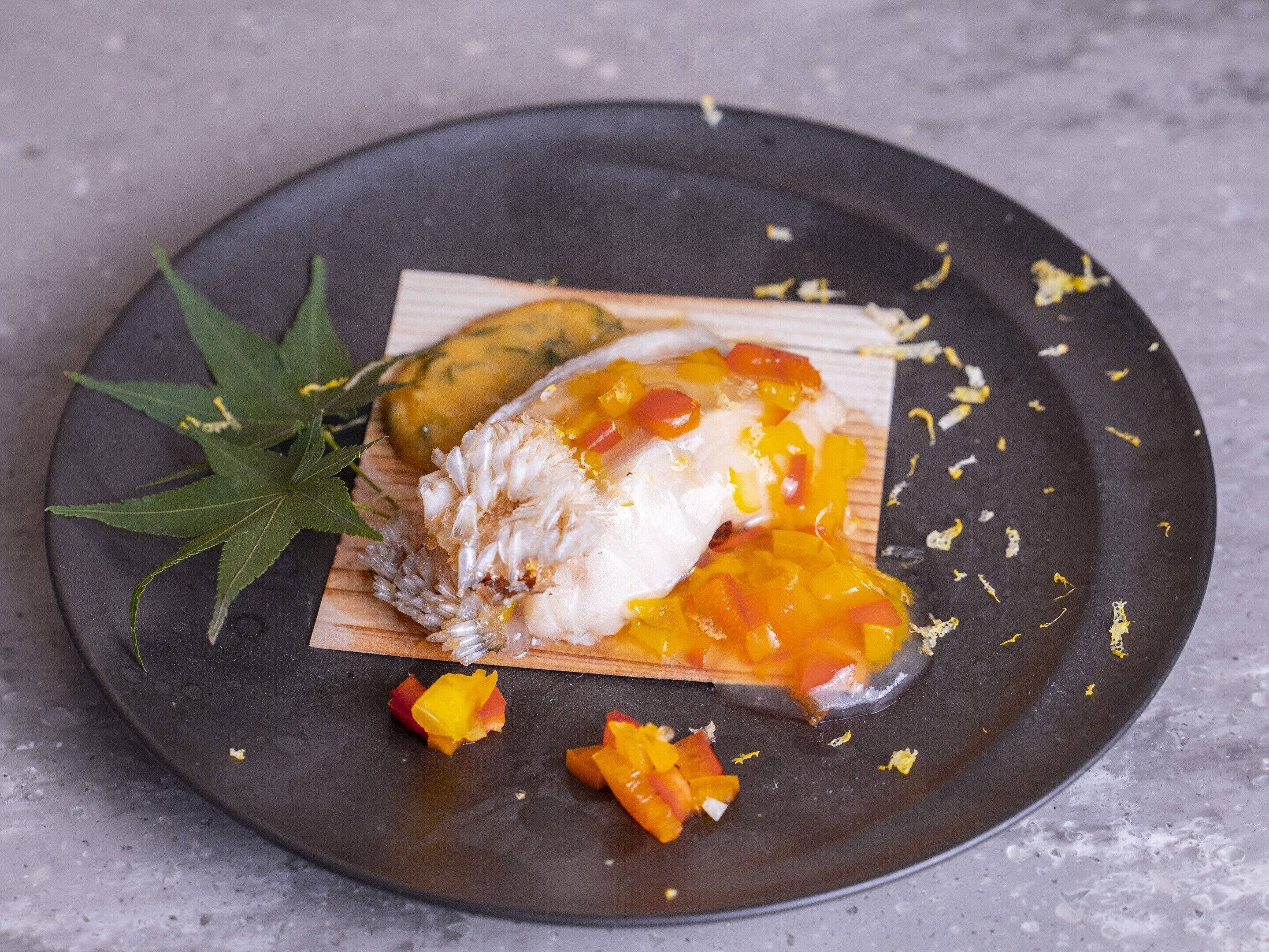 A plate with a white fish fillet topped with diced mango and red bell pepper, garnished with a green leaf, on a dark plate.