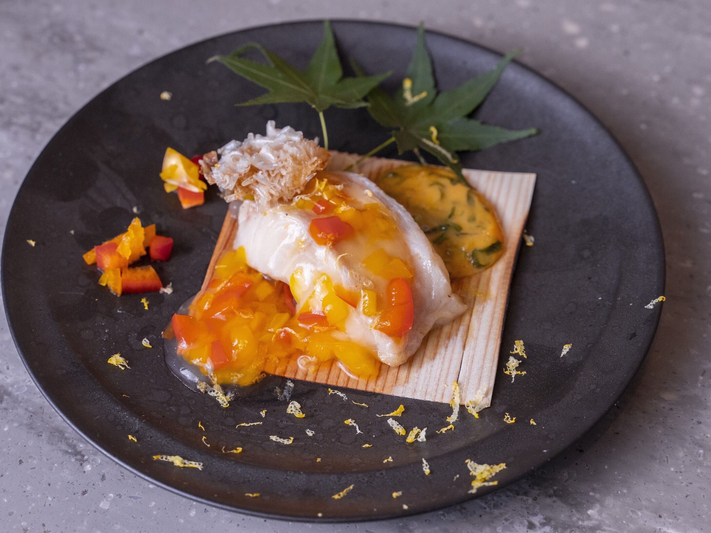 A dish with fish, diced vegetables, and a garnish on a dark plate.