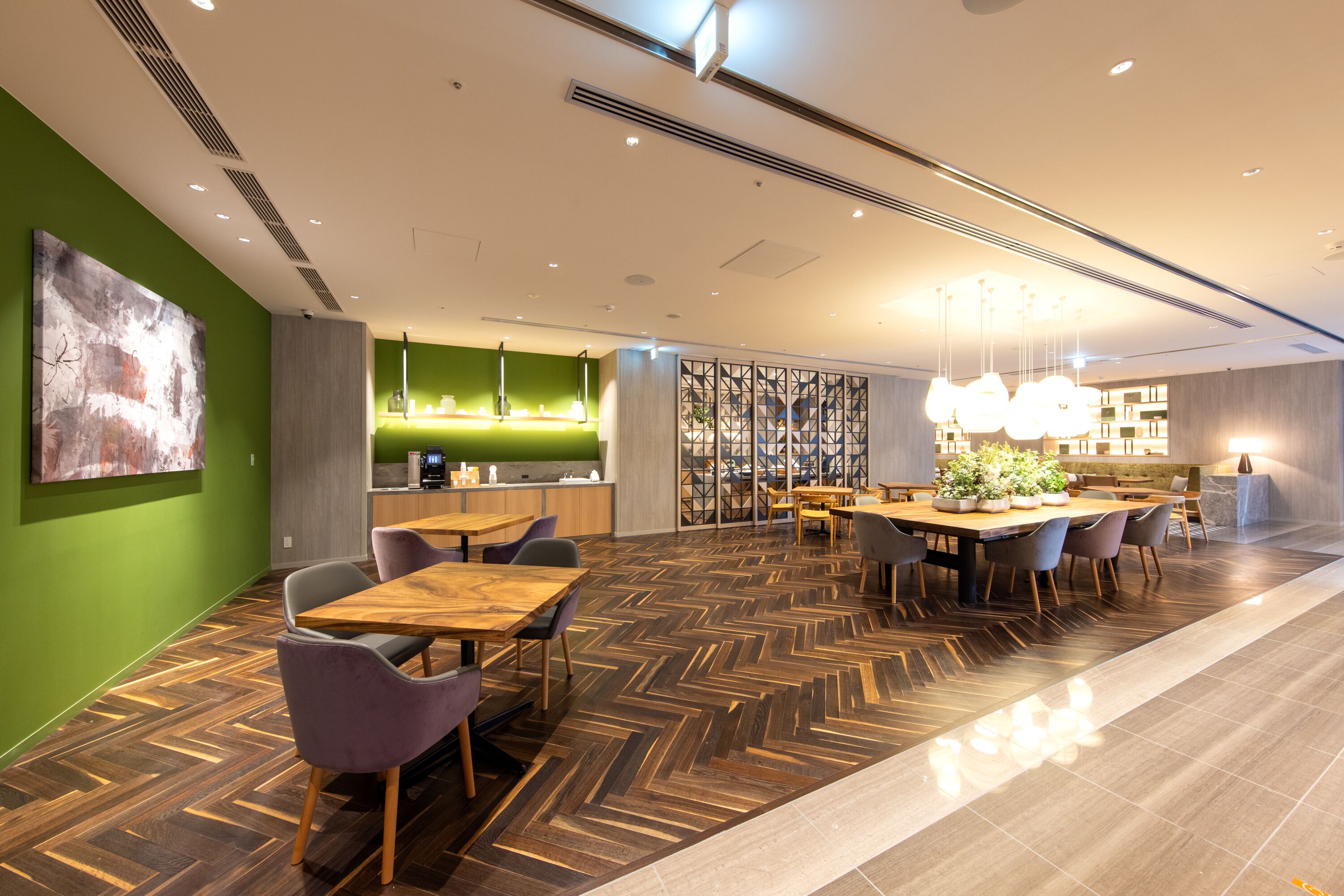 A modern restaurant with wooden tables and chairs, a green accent wall, and a herringbone floor pattern.
