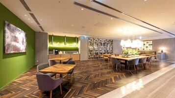 A modern restaurant with wooden tables and chairs, a green accent wall, and a herringbone floor pattern.