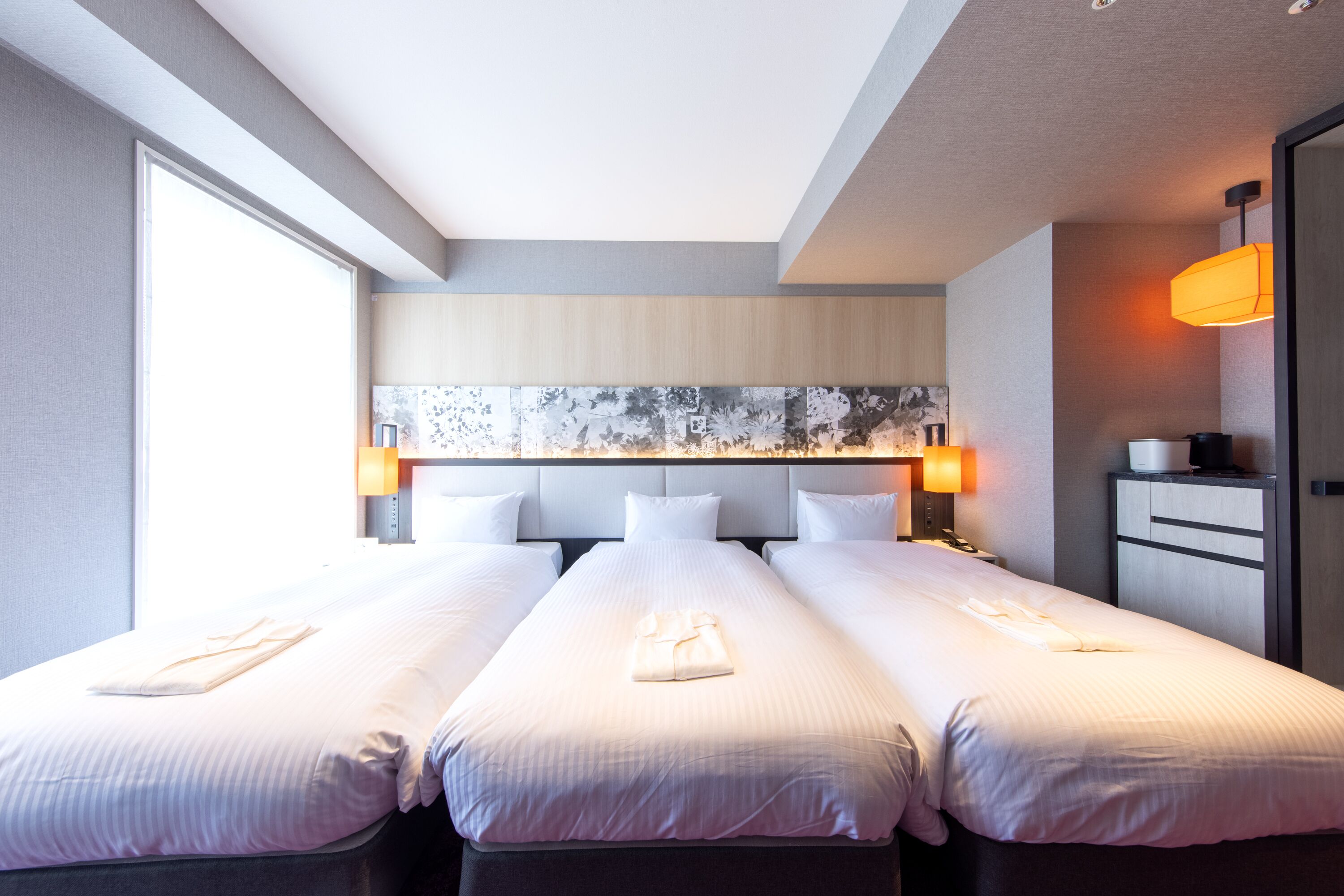Een hotelkamer met twee bedden, een groot raam, een hoofdeinde met een bloemistisch kunstwerk en een nachtkastje met een lamp.