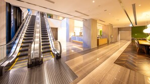 Vue intérieure d’un hall d’hôtel moderne, avec un escalier mécanique menant au deuxième étage, un comptoir d’accueil et un espace repas doté d’un plancher en bois.