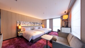 Une chambre d’hôtel avec deux lits, une petite table, un canapé, une grande fenêtre et une œuvre d’art accrochée au mur.