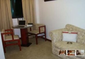 Um quarto pequeno com um sofá bege, uma cadeira de madeira, uma mesinha com um laptop e uma janela com cortinas.