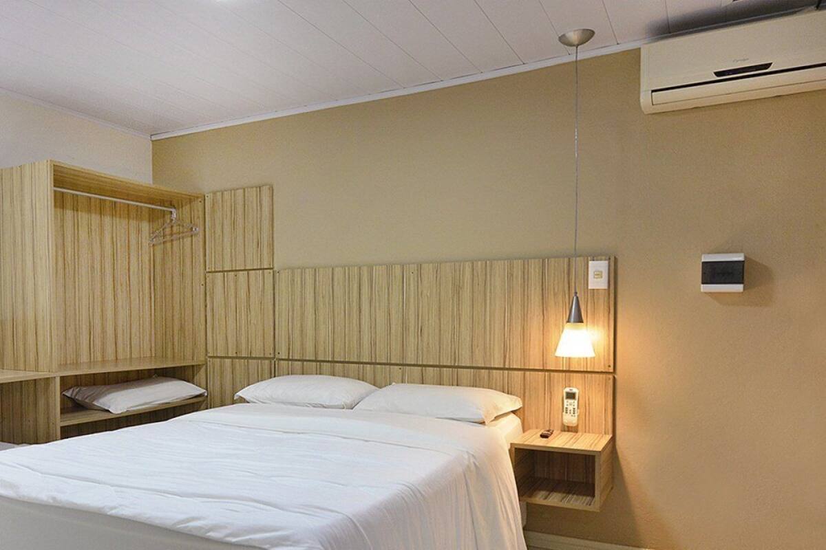 A hotel room with a bed, a bedside table with a lamp, a hanging rack, and a wall-mounted air conditioner.