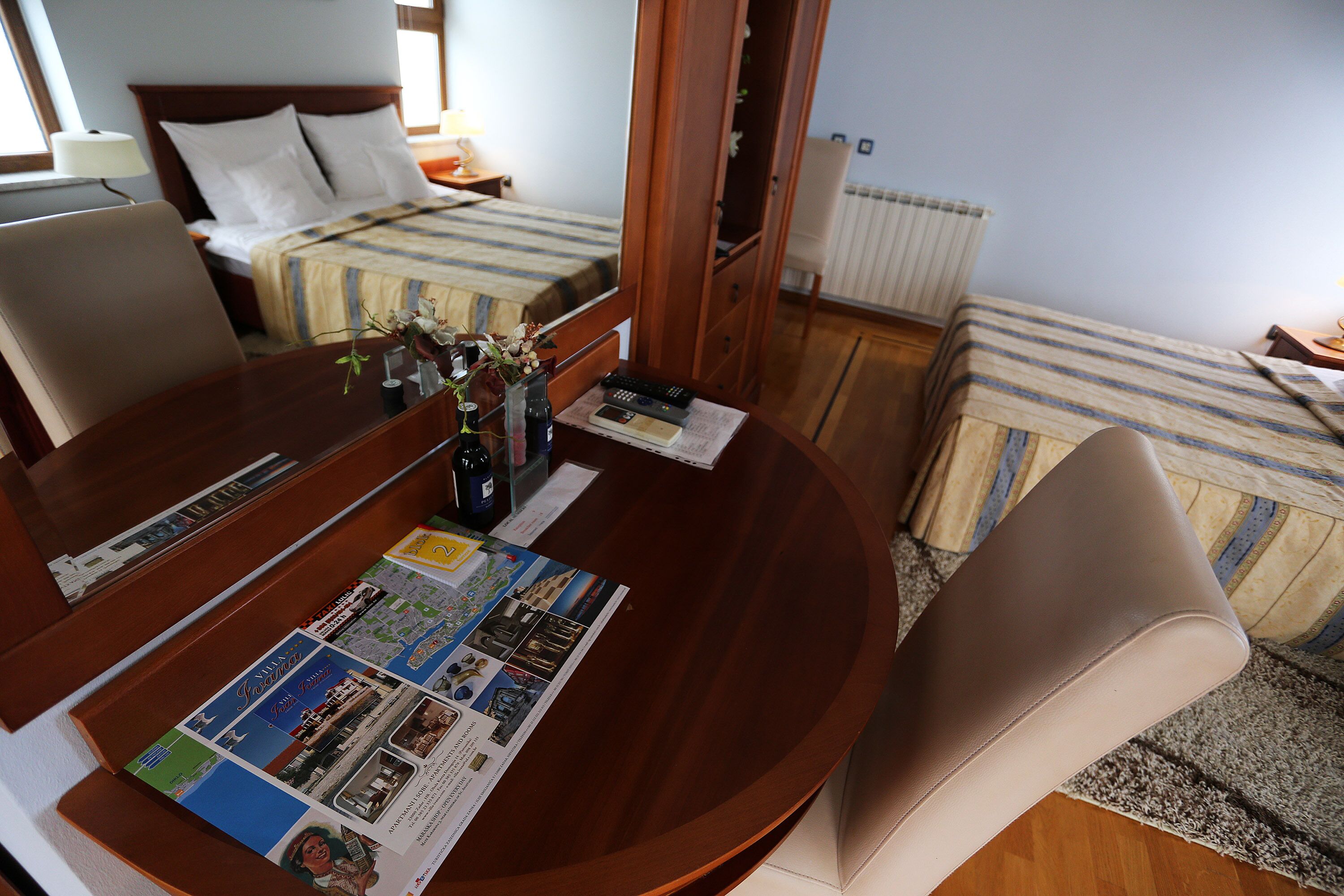 Habitación de hotel con un escritorio de madera, una cama con colcha a rayas y una pequeña mesa con un jarrón de flores y una revista.