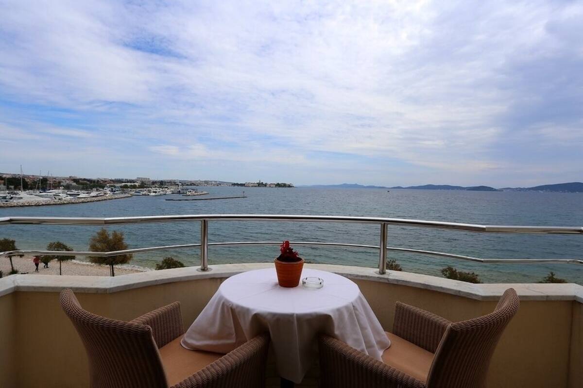 A balcony with wicker chairs and a table, overlooking a marina and the sea.