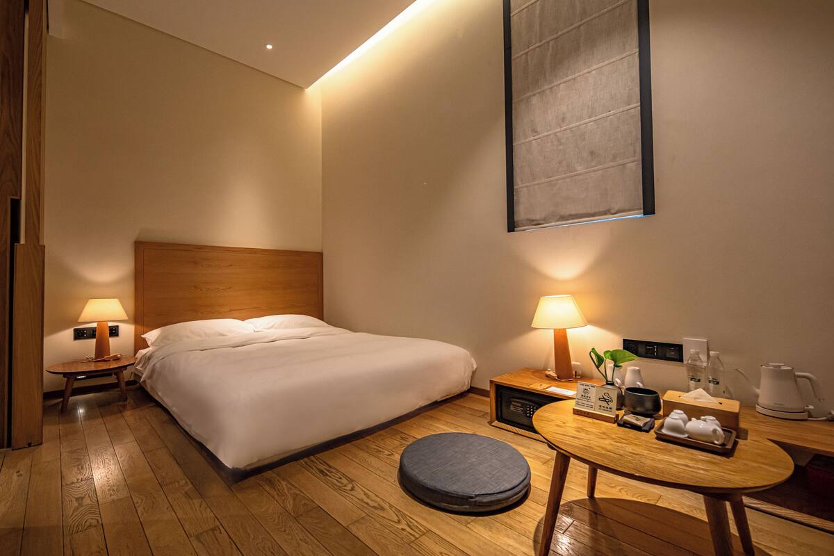 Une chambre d’hôtel comprenant un lit, des tables de chevet avec des lampes, un pouf rond, une table en bois avec des accessoires et un grand tableau abstrait accroché au mur.