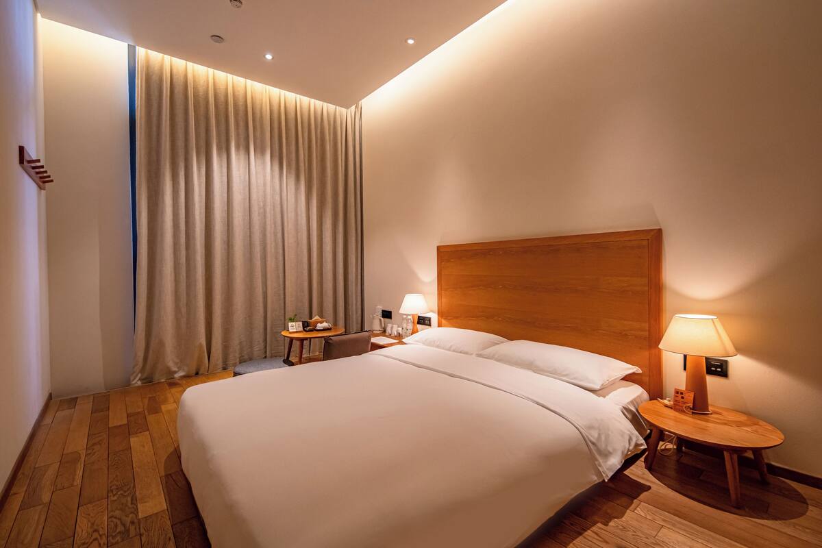 Une chambre d’hôtel avec un grand lit, une tête de lit en bois, une table de chevet avec une lampe, une petite table avec un téléphone et une tasse, et un rideau à la fenêtre.
