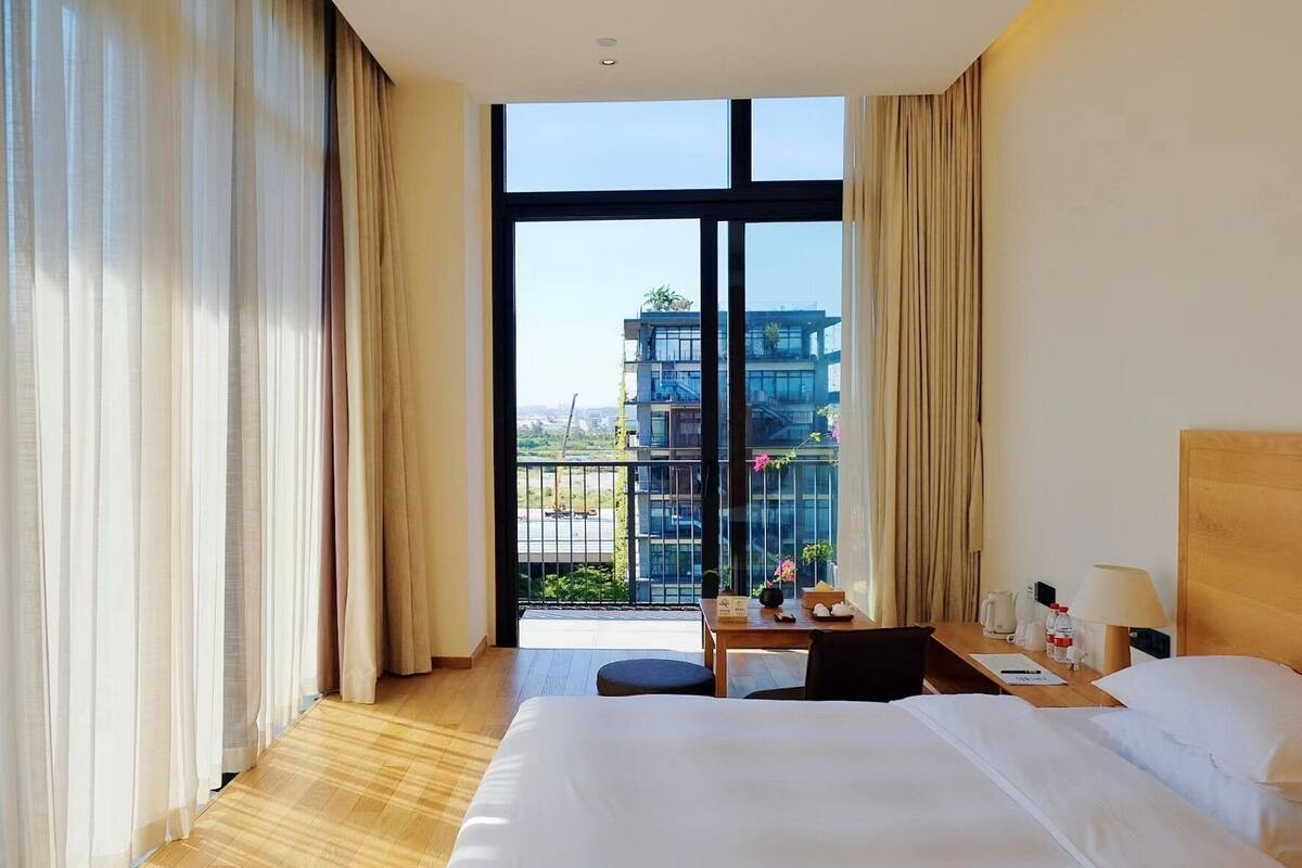 Une chambre d’hôtel avec un grand lit, un bureau avec une lampe et une vue sur les bâtiments, visible à travers de grandes fenêtres.