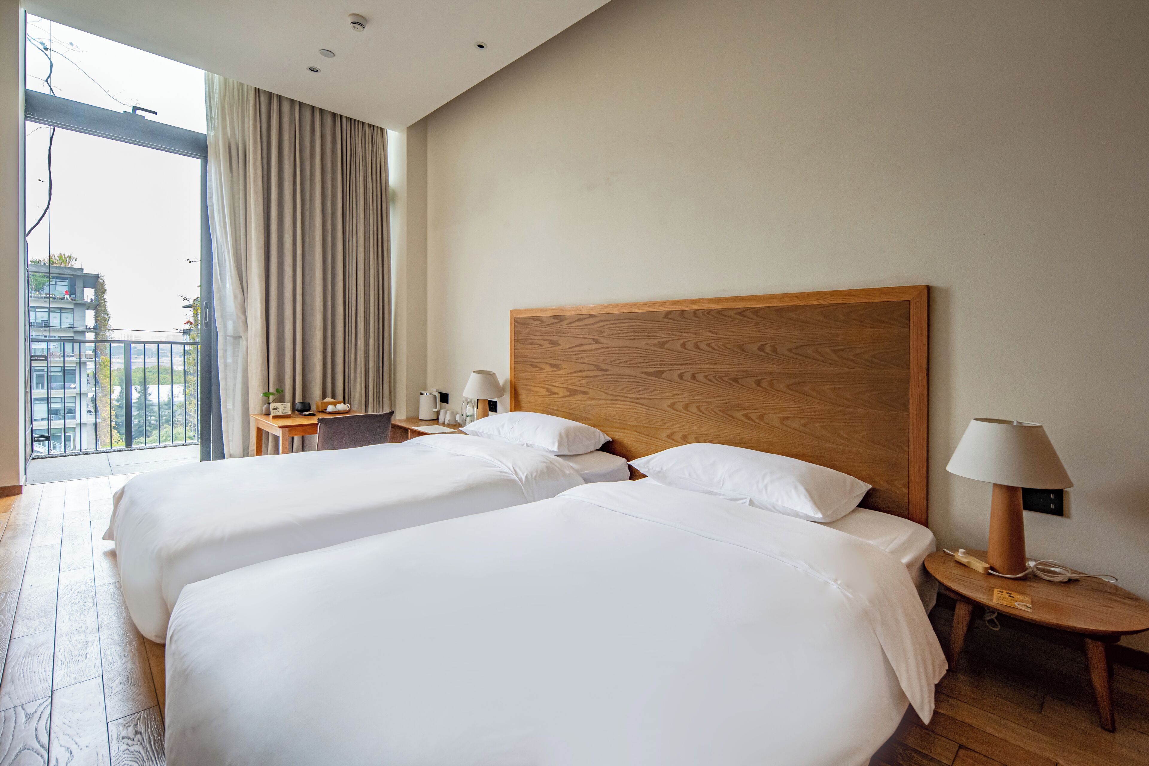 Quarto de hotel com duas camas, cabeceira de madeira, mesa de cabeceira com abajur e vista da cidade por grandes janelas.