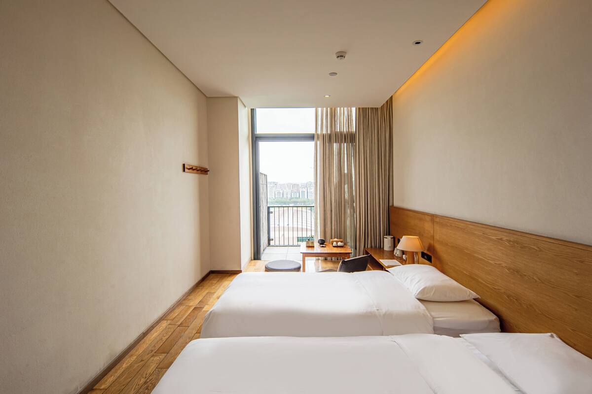 Une chambre d’hôtel avec deux lits, une tête de lit en bois, une petite table avec une lampe et une vue sur la ville par une grande fenêtre.