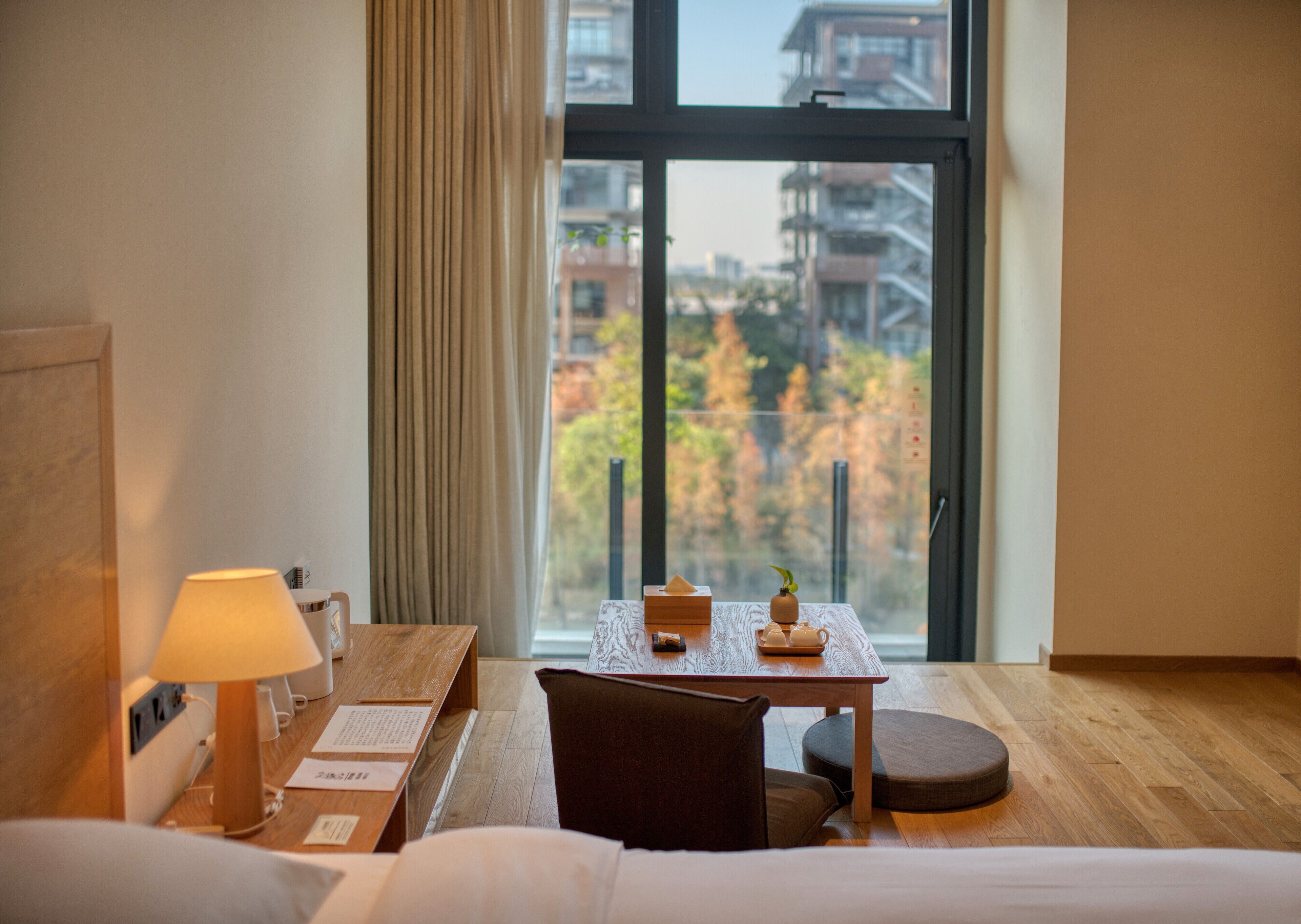 A hotel room with a wooden desk, a lamp, a chair, a small table with a teapot and cups, and a view of autumn trees through a large window.