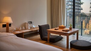 Une chambre d’hôtel avec un lit, un bureau doté d’une lampe, une chaise, une table avec une théière, et une vue sur les bâtiments à travers de grandes fenêtres.
