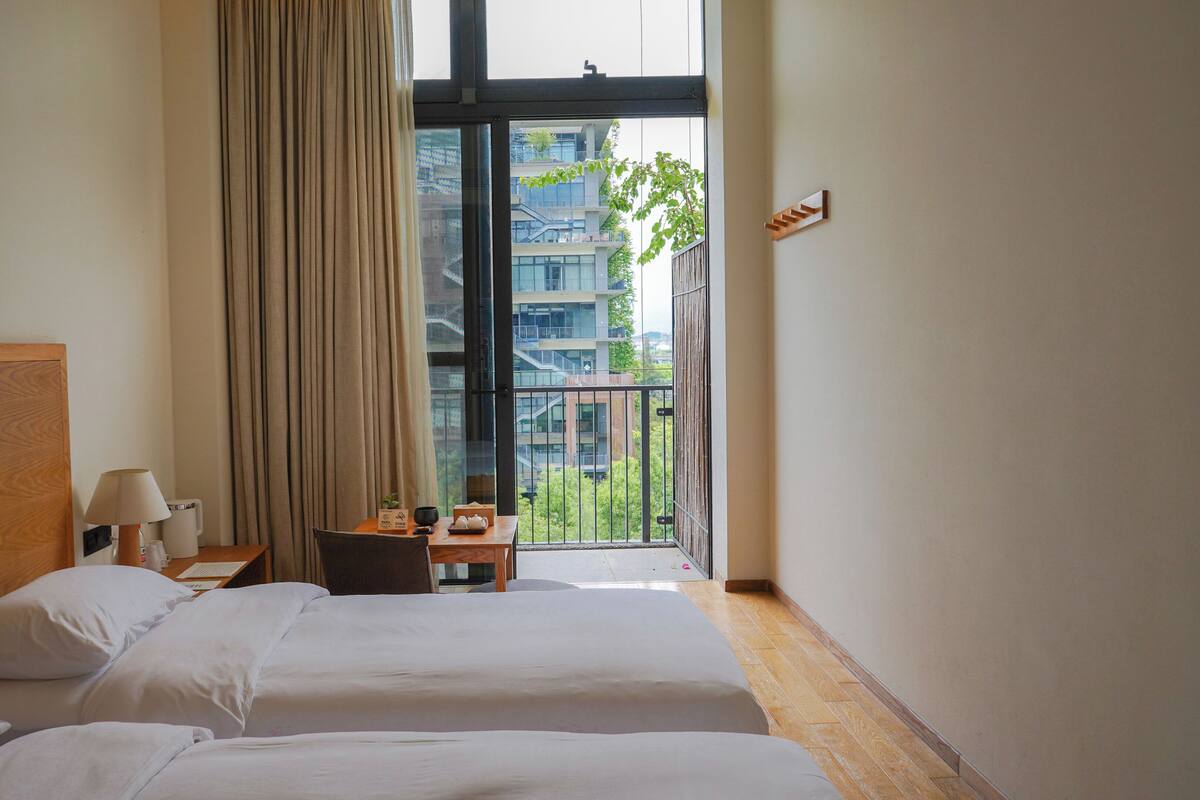 Une chambre d’hôtel avec deux lits, une petite table et une chaise, une fenêtre avec des rideaux, et une vue sur un balcon et des immeubles.