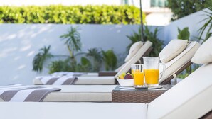 A sunlit outdoor lounge area with reclining chairs, a wicker table with a fruit bowl and glasses of orange juice, and a white wall with greenery in the background. - Holiday home Lana (Supetar)