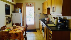 A compact kitchen with wooden cabinets, a refrigerator, a stove, and a coffee maker. There is a dining table with chairs and a window with a floral curtain.